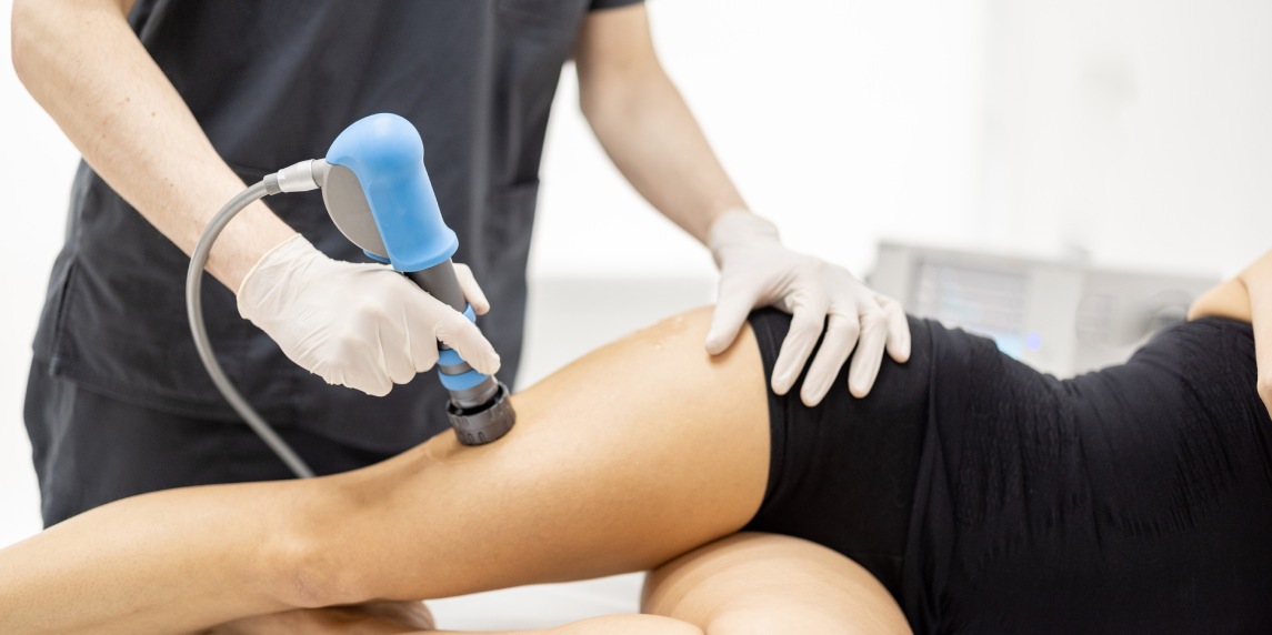 Doctor applies shock wave therapy with special medical equipment on women's thigh muscle at medical office. Concept of non-invasive technology for treating pain in musculoskeletal system