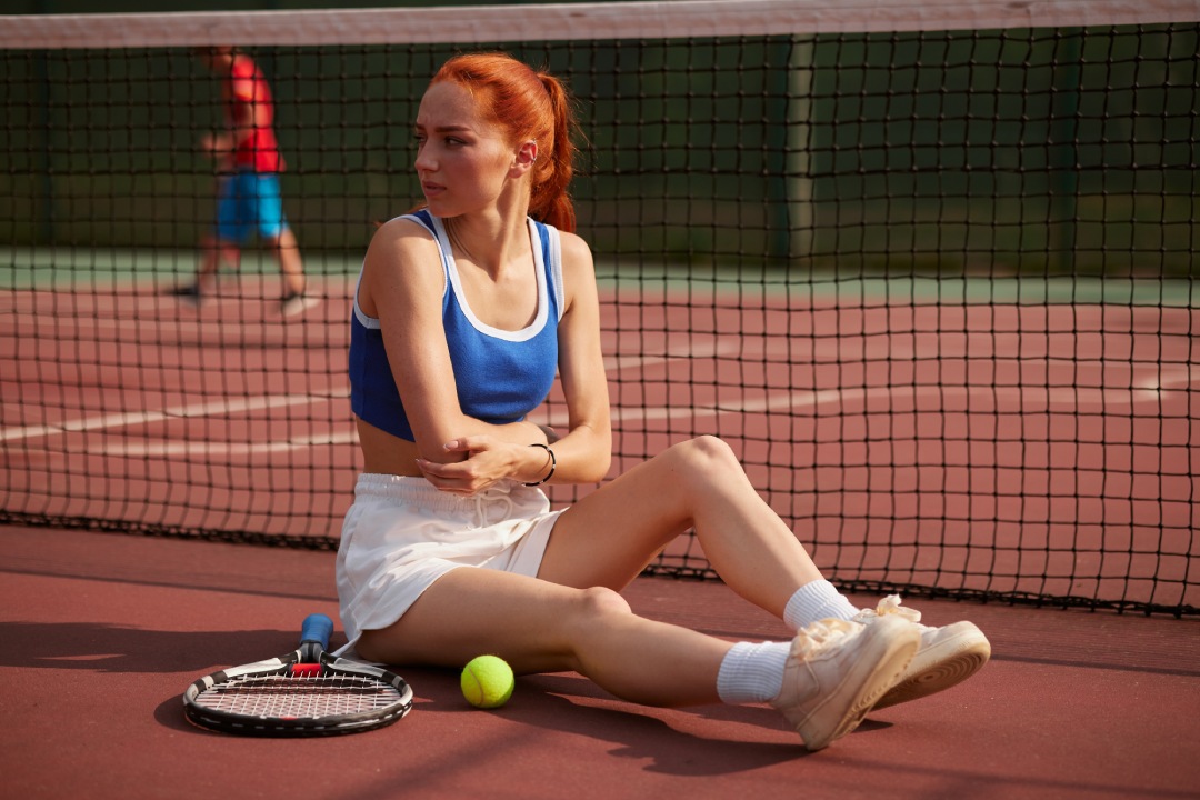 Young female tennis player sitting on court injured knee while playing a match and showing a face of pain