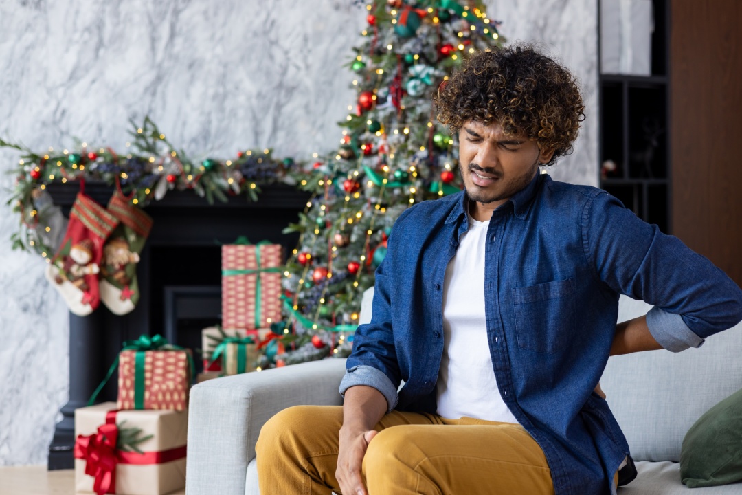 A sick man on Christmas has severe back pain, massages muscle his hand sitting on sofa in living room. Hispanic celebrating New Year and Christmas holidays on a winter day near tree with gifts.